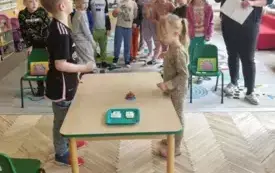 chłopiec i dziewczynka stoją na przeciwko siebie pomiedzy nimi stolik a na nim dzwonek i karteczki w tle pozostałe dzieci i pani nauczycielka