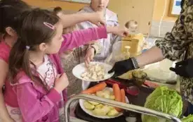 trzy dziewczynki przy w&oacute;zku gastronomicznym wrzucają warzywa do szatkownicy obok nauczycielka