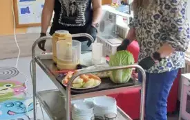 dwie panie przy w&oacute;zku gastronomicznym na kt&oacute;rym znajdują się talerzyki kubeczki warzywa szatkownica naczynia