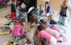 dzieci siedzą na dywanie układają obrazki wśr&oacute;d nich dwie panie jedna z pań podaje obrazki