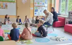 dzieci siedzą w kole na dywanie z nimi dwie panie jedna siedzi na krzesełku i trzyma książkę druga pani siedzi obok na kanapie