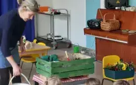 pani nauczycielka trzyma wiadro z ziemią przed nią skrzynka z przyrządami ogrodniczymi i skrzyneczka z bratkami dzieci siedzą na dywanie