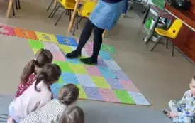 dzieci siedzą na dywanie pani nauczycielka stoi na macie ułożonej z kolorowych cyfr
