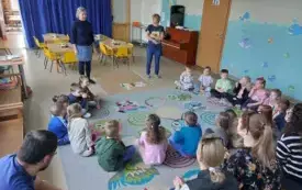 dzieci siedzą na dywanie w p&oacute;łkolu przed nimi stoją dwie panie nauczycielki na podłodze leży obrazek