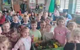 grupa dzieci pozuje do zdjęcia przed nimi skrzynka z kwiatami w tle widać rodzic&oacute;w i nauczycielki