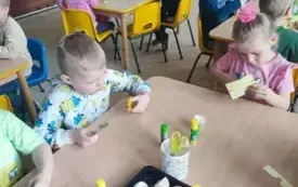 dzieci siedzą przy stolikach na kt&oacute;rych stoją kleje i r&oacute;żne plastyczne elementy oraz skorupki jajek