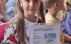 dziewczynka pozuje do zdjęcia pokazując wyr&oacute;żnienie w konkursie w tle inne dzieci