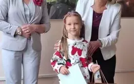 dziewczynka pozuje do zdjęcia trzyma dyplom i torebkę prezentową po bokach stoją dwie elegancko ubrane panie