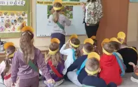 dzieci w ż&oacute;łtych opaskach siedzą przed tablicą na kt&oacute;rej wisi zdjęcie łąki są przyczepione wyrazy obok stoi pani nauczycielka przed tablicą stoi dziewczynka