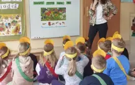 dzieci w ż&oacute;łtych opaskach siedzą przed tablicą na kt&oacute;rej wisi zdjęcie łąki są przyczepione wyrazy obok stoi pani nauczycielka