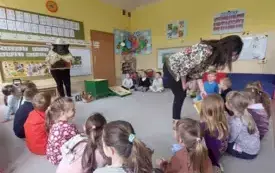 dzieci siedzą w kole pani nauczycielka w stroju pszczelarza prezentuje dzieciom elementy ula kt&oacute;ry stoi przed nią druga pani pokazuje element dzieciom z bliska