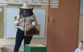 dzieci siedzą w kole pani nauczycielka w stroju pszczelarza prezentuje dzieciom elementy ula kt&oacute;ry stoi przed nią