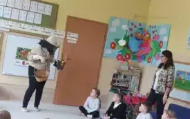 dzieci siedzą w kole dwie panie nauczycielki stoją jedna z pań ubrana w str&oacute;j pszczelarza trzyma w ręku koszyk z akcesoriami