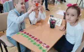 dziewczynka i dw&oacute;ch chłopc&oacute;w w biało-granatowych strojach siedzą przy stoliku na kt&oacute;rym widać akcesoria matematyczne chłopiec pokazuje czerwony element