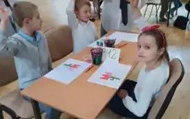 dziewczynka i dw&oacute;ch chłopc&oacute;w w biało-granatowych strojach siedzą przy stoliku na kt&oacute;rym widać akcesoria matematyczne chłopcy mają uniesione po jednej ręce przy nich stoi pochylona pani