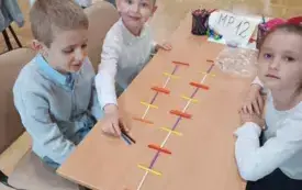 dziewczynka i dw&oacute;ch chłopc&oacute;w w biało-granatowych strojach siedzą przy stoliku na kt&oacute;rym widać akcesoria matematyczne