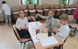 dziewczynka i dw&oacute;ch chłopc&oacute;w w biało-granatowych strojach siedzą przy stoliku na kt&oacute;rym widać akcesoria matematyczne