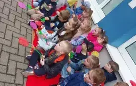 dzieci siedzą na czerwonych kocach pod ścianą trzymają w dłoniach klapki na muchy w kształcie r&oacute;ż