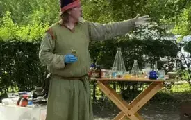 pan alchemik wskazuje na coś ręką za nim st&oacute;ł z przyborami i akcesoriami alchemicznymi