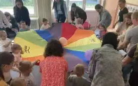 pani nauczycielka wraz z dziećmi i rodzicami trzymają chustę animacyjną na kt&oacute;rej są balony