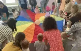 pani nauczycielka wraz z dziećmi i rodzicami siedzą dokoła chusty animacyjnej na kt&oacute;rej podrzucają balony