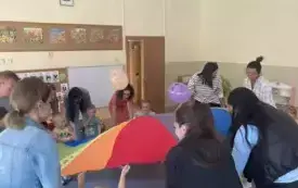 pani nauczycielka wraz z dziećmi i rodzicami siedzą dokoła chusty animacyjnej na kt&oacute;rej podrzucają balony
