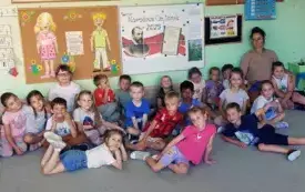 grupa dzieci wraz z nauczycielką pozują do zdjęcia grupowego z tablicą na kt&oacute;rej zawieszony jest plakat promujący narodowe czytanie 2025