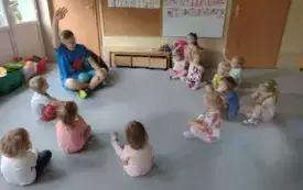 dzieci siedzą na dywanie w kole wśr&oacute;d nich pan ubrany na niebiesko z uniesioną w g&oacute;rze ręką