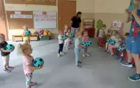 grupa dzieci z niebiesko czarnymi piłkami w rękach stoją na dywanie przed nimi stoi pan z piłką w tym samym kolorze  wśr&oacute;d dzieci pani nauczycielka