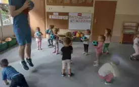 grupa dzieci z niebiesko czarnymi piłkami w rękach stoją na dywanie między nimi pan z piłką w tym samym kolorze uchwycony w skoku nad podłogą