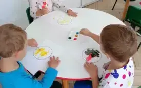 dzieci siedzą przy okrągłym stoliku i stemplują farbami szablony k&oacute;ł