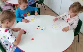 dzieci siedzą przy okrągłym stoliku i stemplują farbami szablony k&oacute;ł