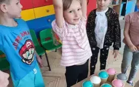 dziewczynka z uniesionymi rękami stoi przed stolikiem obok niej stoi chłopiec i dziewczynka na stole kubeczki w kt&oacute;rych są kolorowe piłeczki