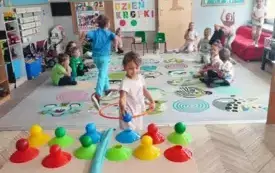 dzieci w klasie kilkoro siedzi na dywanie kilkoro ma założone obręcze na stoliku leżą kolorowe piłeczki na podstawkach