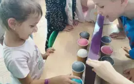 dzieci stoją przy stoliku na kt&oacute;rym stoją kubeczki w nich piłeczki rozdzielone ścianką