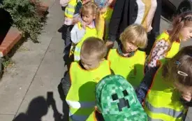 dzieci w parach ubrane w kamizelki odblaskowe wraz z panią stoja na chodniku