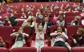 dzieci siedzą na sali teatralnej na czerwonych fotelach  dziewczynka na pierwszym planie unosi do g&oacute;ry ręce