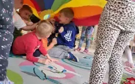 czterech chłopc&oacute;w siedzi pod chustą animacyjną kt&oacute;rą trzymają pozostałe dzieci chłopcy się śmieją