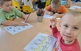 dzieci siedzą przy stolikach na kt&oacute;rych stoją kredki chłopiec patrzy w stronę obiektywu przed nim karta pracy z emplematem korony