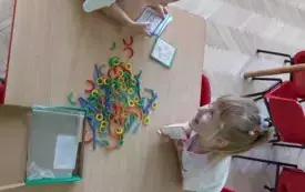 dwie dziewczynki siedzą przy stoliku na kt&oacute;rym leży kolorowa układanka z patyczk&oacute;w w r&oacute;znym kształcie dziewczynka trzyma szablony kształt&oacute;w do ułożenia