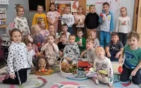 grupa dzieci pozuje do zdjęcia w klasie na podłodze wśr&oacute;d dzieci stoi figurka jeża wykonana z szyszek oraz kosz z darami jesieni