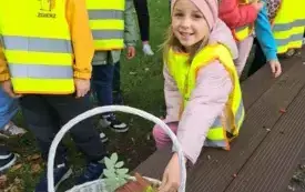 dziewczynka wkłada kolorowy liść do koszyka w tle inne dzieci w kamizelkach odblaskowych