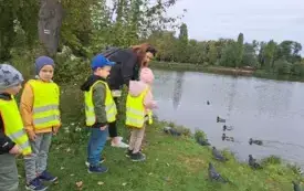 czworo dzieci w kamizelkach odblaskowych stoją z panią nad brzegiem stawu przed nimi gołębie i kaczki