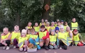 dzieci w kamizelkach odblaskowych pozują do zdjęcia grupowego przedd nimi stoi koszyk z darami jesieni w tle drzewo z tabliczką