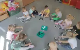 dzieci siedzą w kole na dywanie w środku stoją dwie tace na nich czarne i białe paski bibuły dzieci trzymają paski