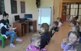 dzieci siedzą wzdł&oacute;ż stołu zwr&oacute;cone w stronę pani kt&oacute;ra siedzi trzymając w dłoniach otwartą książkę