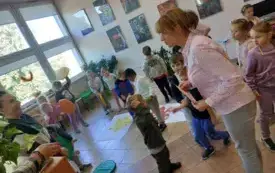 dzieci stoją na sali na pierwszym planie pani nauczycielka trzyma w dłoni kartkę i skierowana jest w stronę drugiej pani