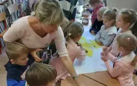 dzieci pochylone nad plakatem z dużym ż&oacute;łtym liściem kt&oacute;ry leży na stole panie stoją nad dziećmi