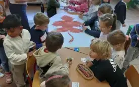 dzieci siedzą przy długim stole na kt&oacute;rym znajdują się kredki i plakat z dużym brązowym liściem obok stoją po przeciwnych stronach stołu dwie panie