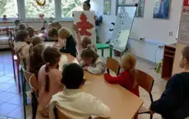 dzieci siedzą przy długim stole na kt&oacute;rym znajdują się kredki i k&oacute;łka z papieru pani trzyma plakat z dużym brązowym liściem